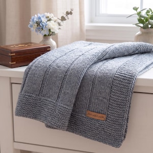 May include: A light blue knitted blanket featuring a textured ribbed pattern and a garter stitch border. A rectangular brown leather tag is attached to the corner, clearly displaying the embossed text "Knit with Love" followed by a small heart symbol.