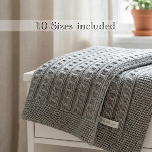 May include: A grey knitted blanket featuring a textured vertical ribbed pattern and a garter stitch border. A small white fabric label with the text "Knit with Love" is attached to the corner. The image includes the text "10 Sizes included".