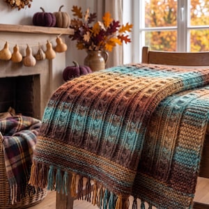 May include: A chunky knit throw blanket featuring a gradient of autumn-inspired colours including deep brown, burnt orange, and teal. The blanket has a textured cable knit pattern and is finished with long, multi-coloured fringe along the bottom edge.