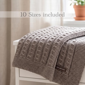 May include: A taupe knitted blanket featuring a textured pattern of vertical columns and a garter stitch border. A small brown rectangular tag with the text "Knit with Love" is attached to the corner. The image includes the text "10 Sizes included".