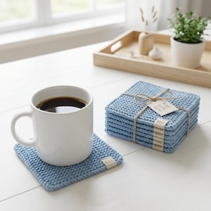 Könnte beinhalten: Eine weiße Tasse mit dunklem Kaffee steht auf einem hellblauen gestrickten Untersetzer. Daneben befindet sich ein Stapel passender Untersetzer, mit Bindfaden und einem Etikett mit der Aufschrift "Knit with Love". Die quadratischen Untersetzer sind strukturiert.