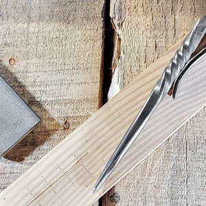 May include: A silver metal tool with a twisted handle and a sharp point. The tool is lying on a wooden surface.