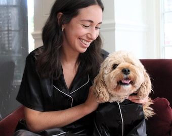 May include: Black satin pajama set with white piping, worn by a person and a dog. The person is smiling while holding the dog, who is also wearing a matching pajama top. The set includes a short-sleeved top and shorts.