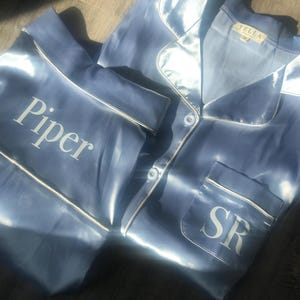 May include: Light blue satin pajama set with white piping. The top has a pocket with the initials "SR" and the word "Piper" is printed on the pants. The tag reads "TELLA".
