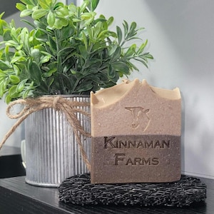 May include: A rectangular bar of two-toned brown soap featuring an embossed goat head icon and the text Kinnaman Farms. The soap rests on a black textured soap dish, positioned in front of a silver corrugated metal planter with a green plant.