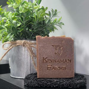 May include: A rectangular brown bar of soap with the embossed text Kinnaman Farms and a goat logo. The soap rests on a black textured soap dish, positioned in front of a small green plant in a corrugated metal pot tied with a brown twine bow.