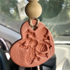 May include: A terracotta heart-shaped car air freshener with a bee design. The air freshener is hanging from a string with two wooden beads.