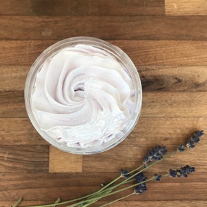 May include: A round, clear plastic container filled with light purple whipped body butter, featuring a swirled, piped texture on the surface. Several sprigs of dried lavender with small purple flower buds and green stems are positioned nearby.