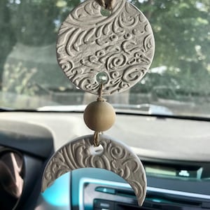 May include: A cream-colored car charm featuring a circular pendant and a crescent moon pendant, both with an embossed floral design. The charm includes wooden beads and is suspended by a natural-colored cord. The background shows a car interior and a blurred view of trees.