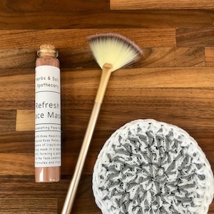 May include: A glass bottle with a cork stopper containing a pink face mask labeled "Refresh Face Mask" with the text "Herbs & Soil Apothecary" and "Rejuvenating Face Mask" on the label. A gold fan brush and a white and gray crocheted round pad are also in the image.