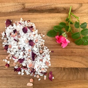May include: A pile of white bath salts with pink Himalayan salt and dried rose petals on a wooden surface. A single pink rose with green leaves is on the right side of the image.