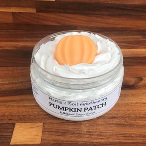 May include: A white jar with a white whipped sugar scrub inside. The jar has a label that reads "Herbs & Soil Apothecary Pumpkin Patch Whipped Sugar Scrub". A small orange pumpkin-shaped soap is on top of the scrub.