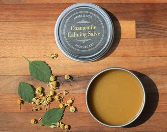 May include: A silver tin of chamomile calming salve with a white label that reads "Herbs & Soil Chamomile Calming Salve Apothecary". The tin is open and the salve is a light yellow color. Dried chamomile flowers and green leaves are scattered on a wooden surface.