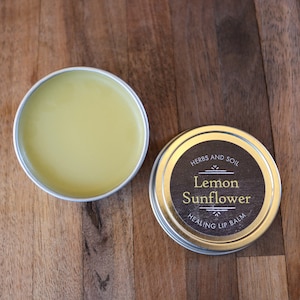 May include: Two silver tins of lip balm, one open and one closed. The open tin reveals yellow balm. The closed tin has a black label with gold text: "Lemon Sunflower Healing Lip Balm" and "Herbs and Soil".
