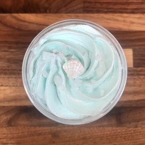 May include: A clear glass jar filled with light blue whipped bath soap. A small white seashell is on top of the soap.