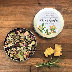 May include: A metal tin of "Herbal Garden Loose Incense" with a floral label, open to reveal a mix of dried herbs and flowers. Beside it are pieces of ginger and a sprig of rosemary on a wooden surface.