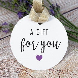 May include: A white round gift tag with a gold ribbon attached. The tag has black text that says "A Gift for You" and a small purple heart.