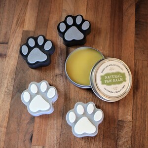 May include: A tin of natural paw balm with a paw print design on the lid. The balm is handmade and cruelty-free. There are four paw print shaped soaps in the image, two are black with a grey paw print and two are grey with a white paw print.