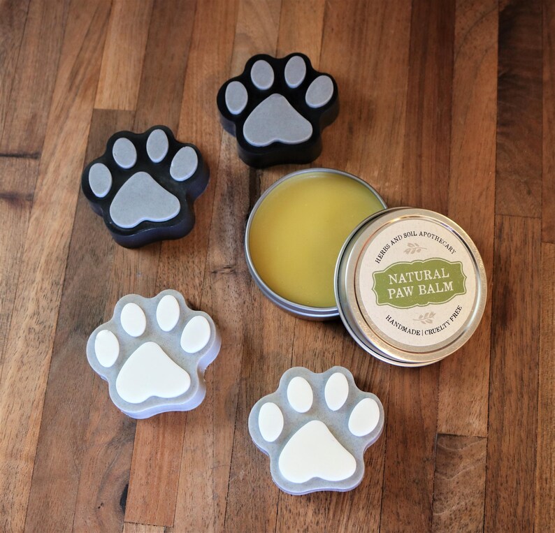 May include: A tin of natural paw balm with a paw print design on the lid. The balm is handmade and cruelty-free. There are four paw print shaped soaps in the image, two are black with a grey paw print and two are grey with a white paw print.