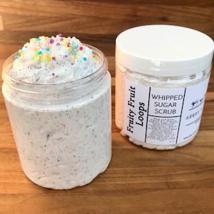 May include: Two jars of whipped sugar scrub, one with a white lid and the other with a clear lid. The jar with the clear lid has a white whipped sugar scrub with colorful sprinkles on top. The jar with the white lid has a label that says "Fruity Fruit Loops Whipped Sugar Scrub".