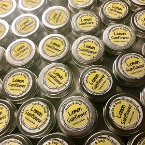 May include: A collection of small, round, silver tins, each with a yellow label. The labels read "Herbs & Soil Lemon Sunflower Healing Lip Balm" in a circular design. The tins are arranged closely together, with the labels facing upwards.