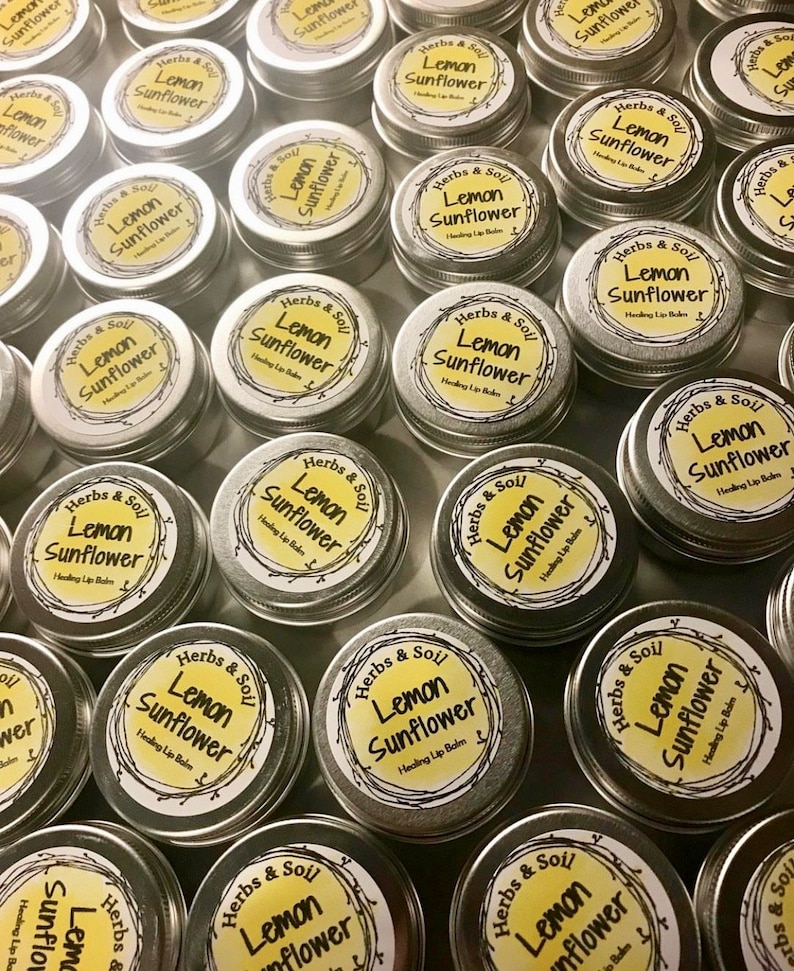 May include: A collection of small, round, silver tins, each with a yellow label. The labels read "Herbs & Soil Lemon Sunflower Healing Lip Balm" in a circular design. The tins are arranged closely together, with the labels facing upwards.