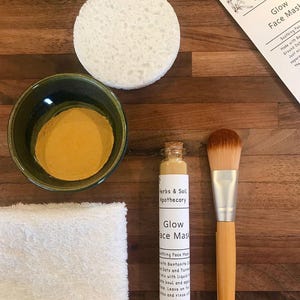 Pode incluir: Um conjunto de produtos naturais para o cuidado da pele para uma m&aacute;scara facial de brilho. O conjunto inclui um pequeno frasco de vidro com p&oacute;, uma pequena garrafa de vidro com rolha de corti&ccedil;a, uma toalha branca, uma tigela verde, uma esponja branca e um pincel de maquiagem de bambu.