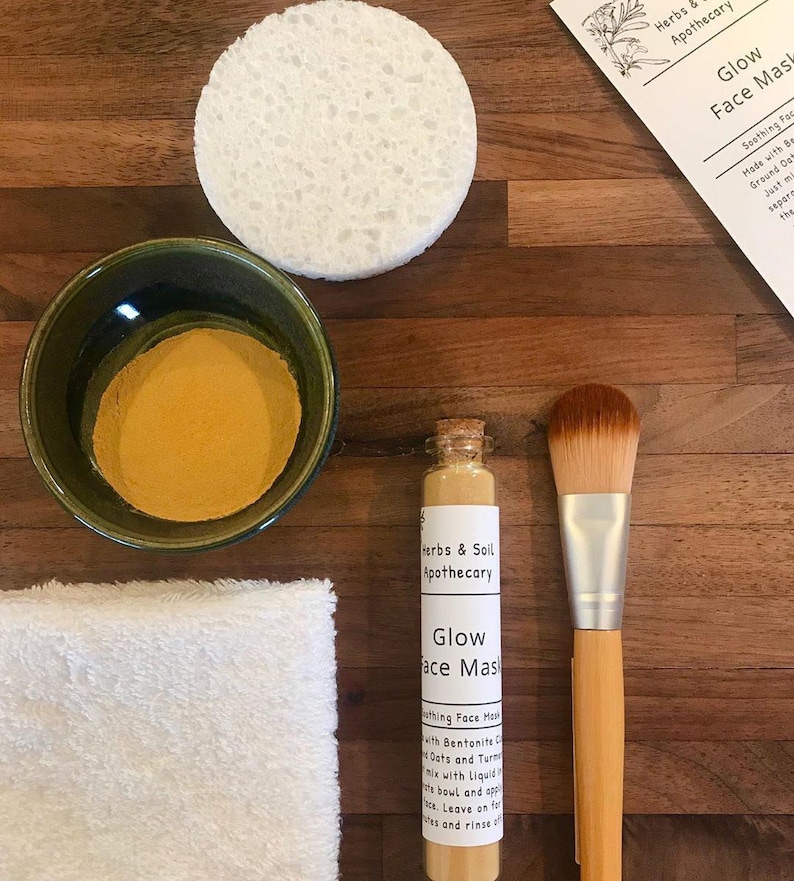Pode incluir: Um conjunto de produtos naturais para o cuidado da pele para uma m&aacute;scara facial de brilho. O conjunto inclui um pequeno frasco de vidro com p&oacute;, uma pequena garrafa de vidro com rolha de corti&ccedil;a, uma toalha branca, uma tigela verde, uma esponja branca e um pincel de maquiagem de bambu.
