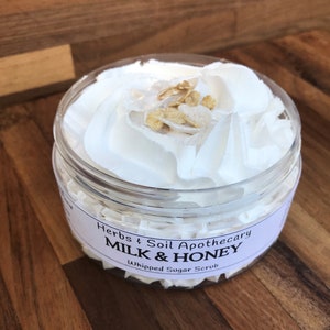 May include: A clear plastic jar filled with white whipped sugar scrub, topped with a swirl of whipped cream and a sprinkle of golden grains. The jar has a label that reads "Herbs & Soil Apothecary MILK & HONEY Whipped Sugar Scrub."
