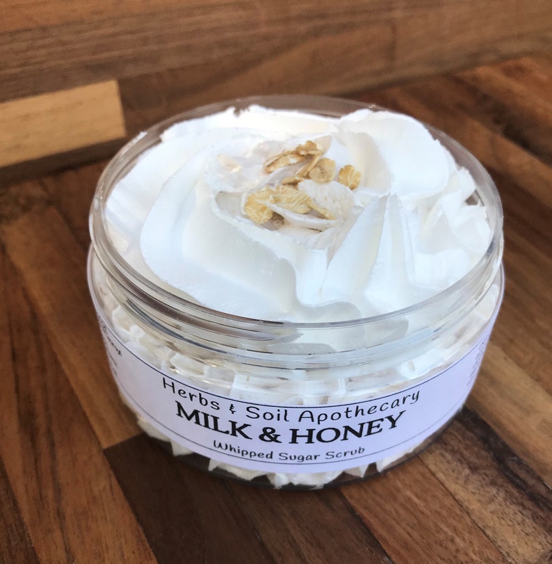 May include: A clear plastic jar filled with white whipped sugar scrub, topped with a swirl of whipped cream and a sprinkle of golden grains. The jar has a label that reads "Herbs & Soil Apothecary MILK & HONEY Whipped Sugar Scrub."