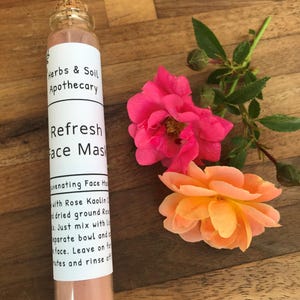 May include: A glass bottle of "Refresh Face Mask" from Herbs & Soil Apothecary, with a cork stopper, next to pink and orange roses. The label lists ingredients and instructions. The mask appears to be a pink powder.
