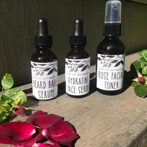 May include: Three amber glass bottles with black caps and white labels. The labels feature black botanical illustrations and text reading "Herbs and Soil Apothecary" followed by "BEARD BABE SERUM", "HYDRATING FACE SERUM", and "ROSE FACIAL TONER".