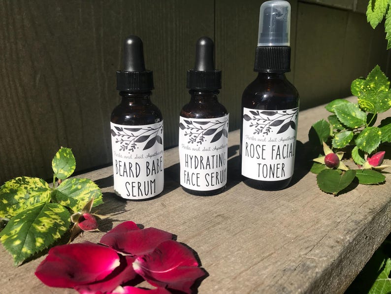 May include: Three amber glass bottles with black caps and white labels. The labels feature black botanical illustrations and text reading "Herbs and Soil Apothecary" followed by "BEARD BABE SERUM", "HYDRATING FACE SERUM", and "ROSE FACIAL TONER".