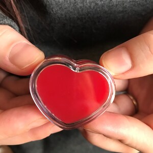 May include: A clear, heart-shaped object with a vibrant red interior. The object is held in open hands against a grey background. The heart shape is outlined with a clear border.