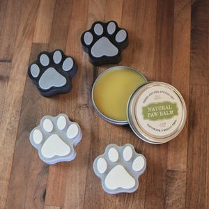 May include: A round metal tin of Natural Paw Balm by Herbs and Soil Apothecary with a yellow waxy substance inside, accompanied by four paw-shaped objects in black and grey or white and grey, featuring a central pad and four smaller toe pads.