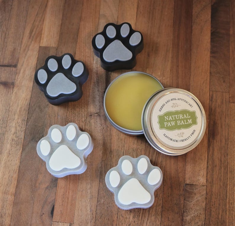 May include: A round metal tin of Natural Paw Balm by Herbs and Soil Apothecary with a yellow waxy substance inside, accompanied by four paw-shaped objects in black and grey or white and grey, featuring a central pad and four smaller toe pads.