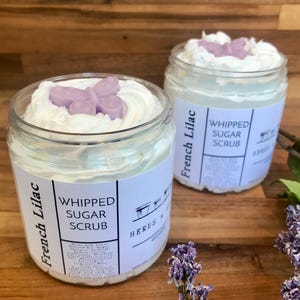 May include: Two clear plastic jars of French Lilac whipped sugar scrub. Each jar features a white label with black text reading French Lilac Whipped Sugar Scrub and Herbs & Soil. The white scrub is topped with a small, light purple butterfly.