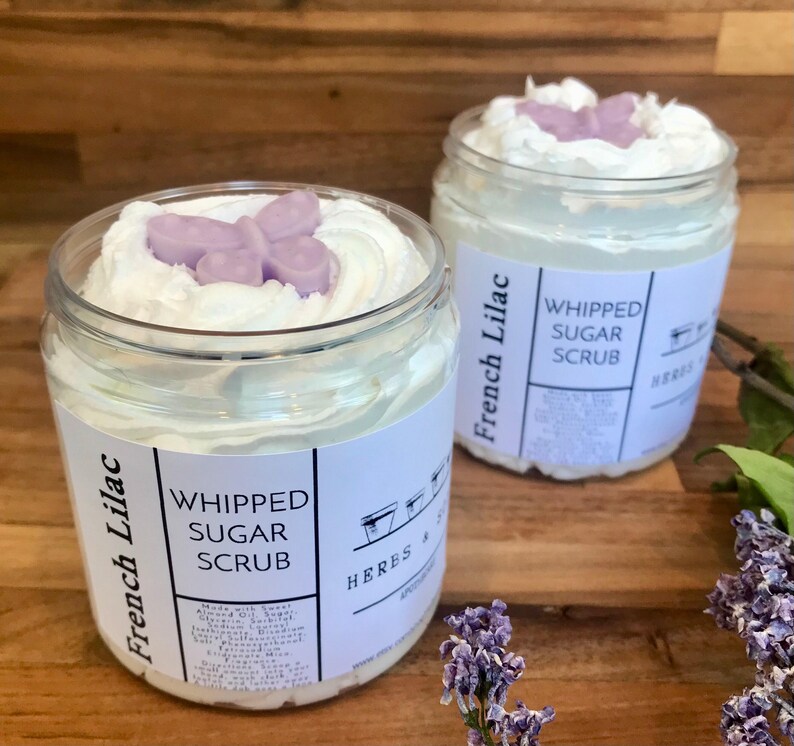 May include: Two clear plastic jars of French Lilac whipped sugar scrub. Each jar features a white label with black text reading French Lilac Whipped Sugar Scrub and Herbs & Soil. The white scrub is topped with a small, light purple butterfly.