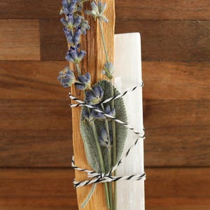 May include: A bundle consisting of a light brown wooden stick, a white crystalline stone, a green sage leaf, and a sprig of purple lavender flowers. These items are secured together with a black and white twisted string wrapped around them.