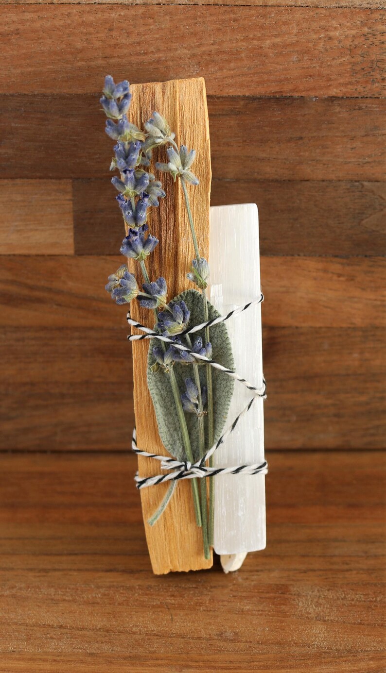 May include: A bundle consisting of a light brown wooden stick, a white crystalline stone, a green sage leaf, and a sprig of purple lavender flowers. These items are secured together with a black and white twisted string wrapped around them.