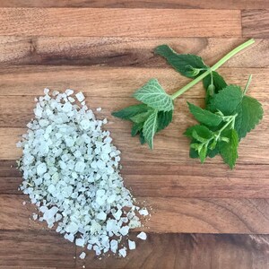 May include: A close-up of coarse, pale salt crystals with green specks, alongside fresh green mint sprigs on a wooden surface. The salt may be herb-infused, suitable for cooking or spa treatments.