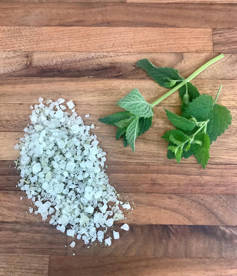 May include: A close-up of coarse, pale salt crystals with green specks, alongside fresh green mint sprigs on a wooden surface. The salt may be herb-infused, suitable for cooking or spa treatments.