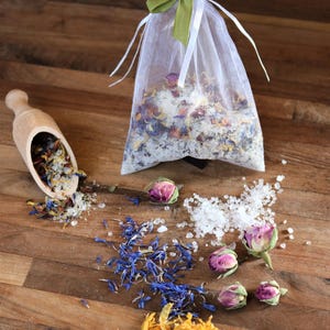May include: A white organza bag filled with bath salts and dried flowers, including rose buds, blue petals, and yellow petals. The bag is tied with a green ribbon. A wooden scoop with bath salts and dried flowers is on the left. There are also loose bath salts and dried flower petals scattered on a wooden surface.