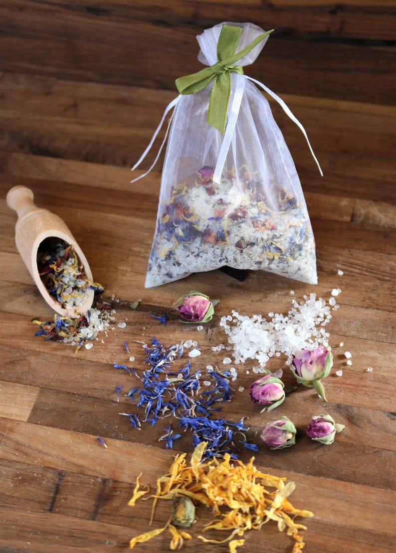 May include: A white organza bag filled with bath salts and dried flowers, including rose buds, blue petals, and yellow petals. The bag is tied with a green ribbon. A wooden scoop with bath salts and dried flowers is on the left. There are also loose bath salts and dried flower petals scattered on a wooden surface.