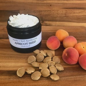 May include: A black jar of Herbs & Soil Apothecary Apricot Seed facial scrub with a white label, filled with a thick white cream. Beside the jar are several whole orange and pink apricots and a pile of light brown, oval-shaped apricot pits.