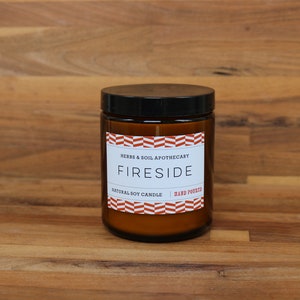 May include: A brown glass candle jar with a black lid. The label reads "Herbs & Soil Apothecary Fireside Natural Soy Candle Hand Poured" with a red and white chevron pattern.