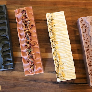 May include: Four rectangular bars of handcrafted soap arranged vertically. The first is charcoal gray with gold dust, the second is dusty rose topped with dried flower petals, the third is cream with botanical bits, and the fourth is mauve with dark specks.