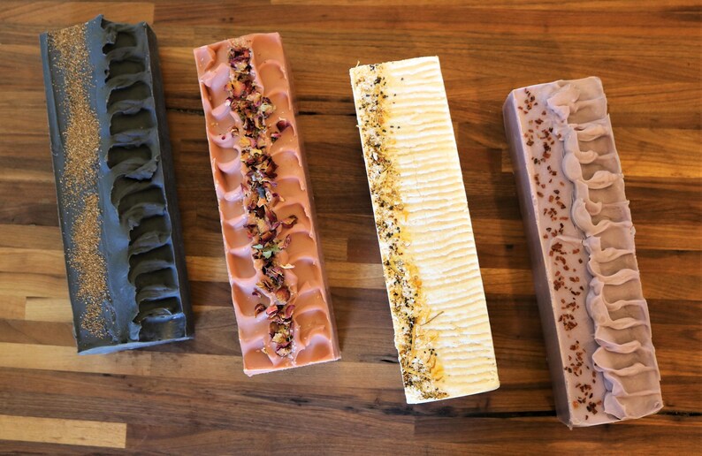May include: Four rectangular bars of handcrafted soap arranged vertically. The first is charcoal gray with gold dust, the second is dusty rose topped with dried flower petals, the third is cream with botanical bits, and the fourth is mauve with dark specks.