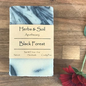 May include: A handmade soap bar with a swirling black and white pattern. The soap is wrapped with a brown paper label that reads "Herbs & Soil Apothecary Black Forest." The net weight is 113-142 grams. The soap is labeled as natural and cruelty-free.