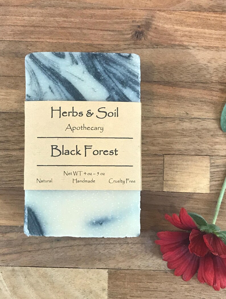May include: A handmade soap bar with a swirling black and white pattern. The soap is wrapped with a brown paper label that reads "Herbs & Soil Apothecary Black Forest." The net weight is 113-142 grams. The soap is labeled as natural and cruelty-free.